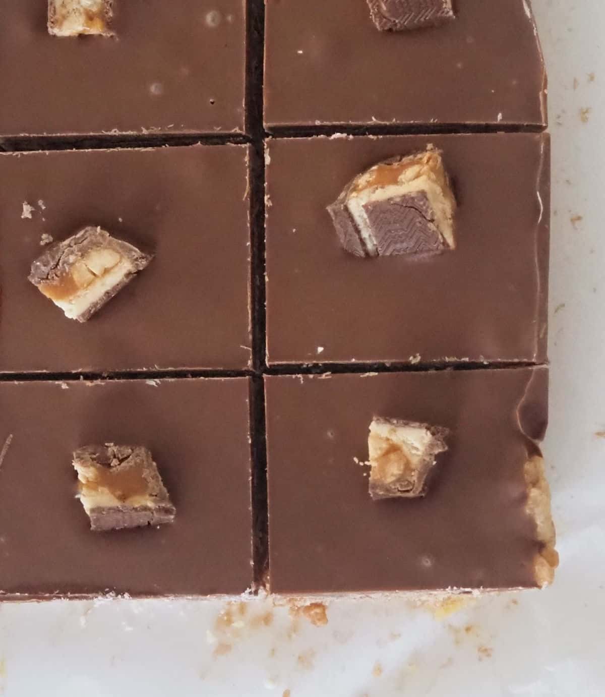 overhead view of 6 pieces of snickers slice sitting on a piece of baking paper. Each piece is topped with a small piece of snickers bar.