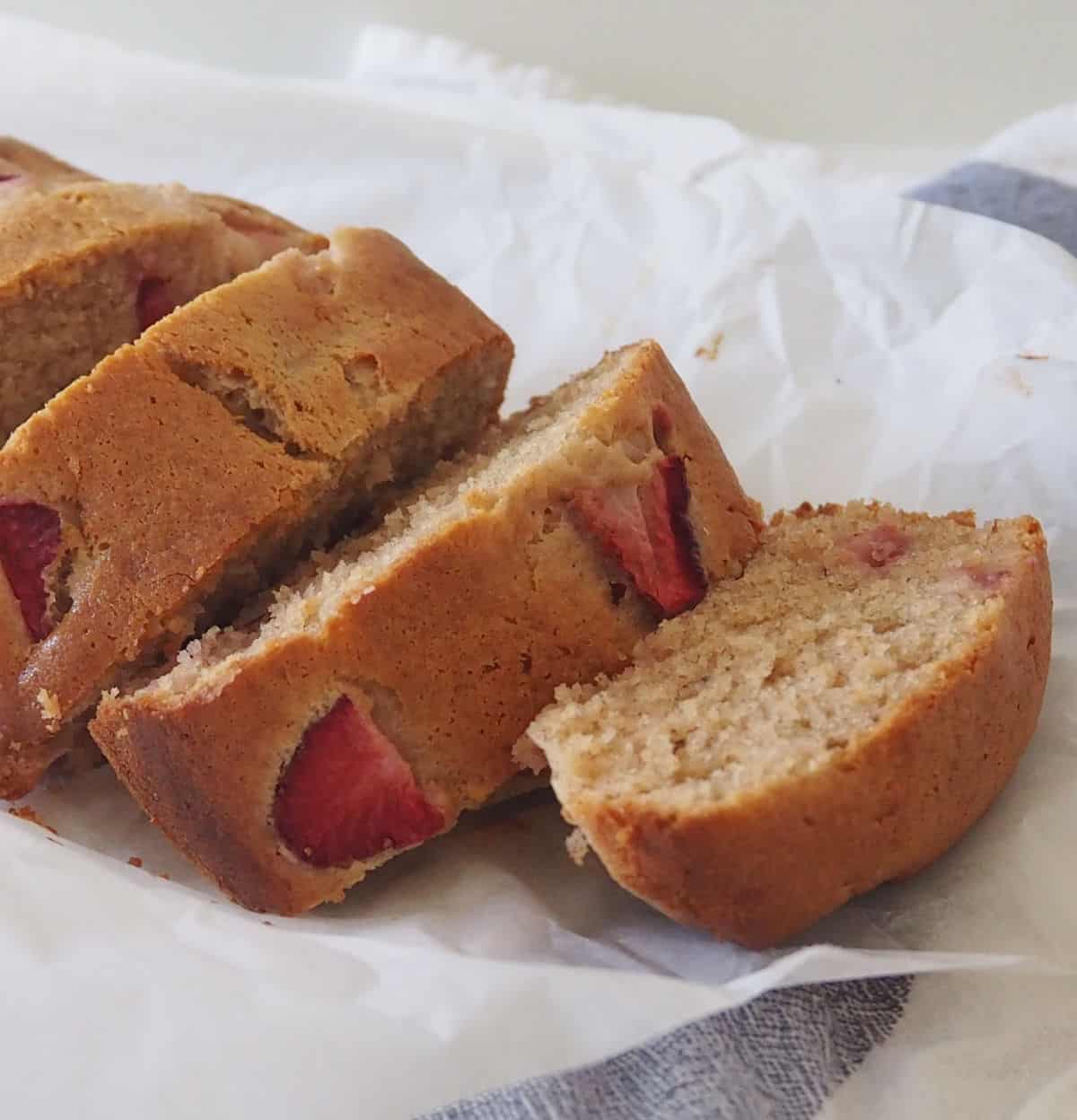 Slices of Banana and Strawberry Loaf sitting on a sheet of baking paper which is on top of a blue check tea towel.