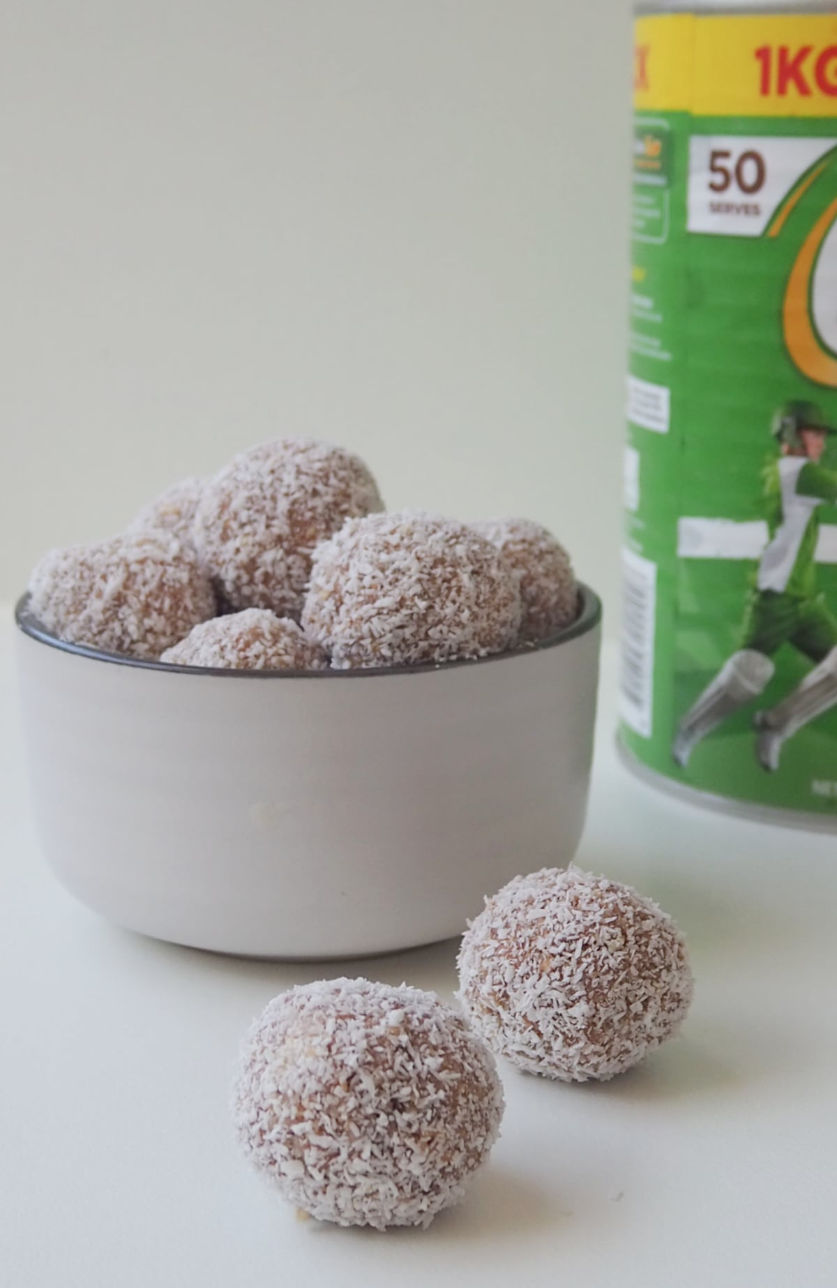 Two Milo Balls on a white surface. In the background is a white bowl with more Milo Balls and a tin of Milo.