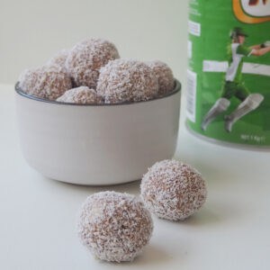 Two Milo Balls on a white surface. In the background is a white bowl with more Milo Balls and a tin of Milo.