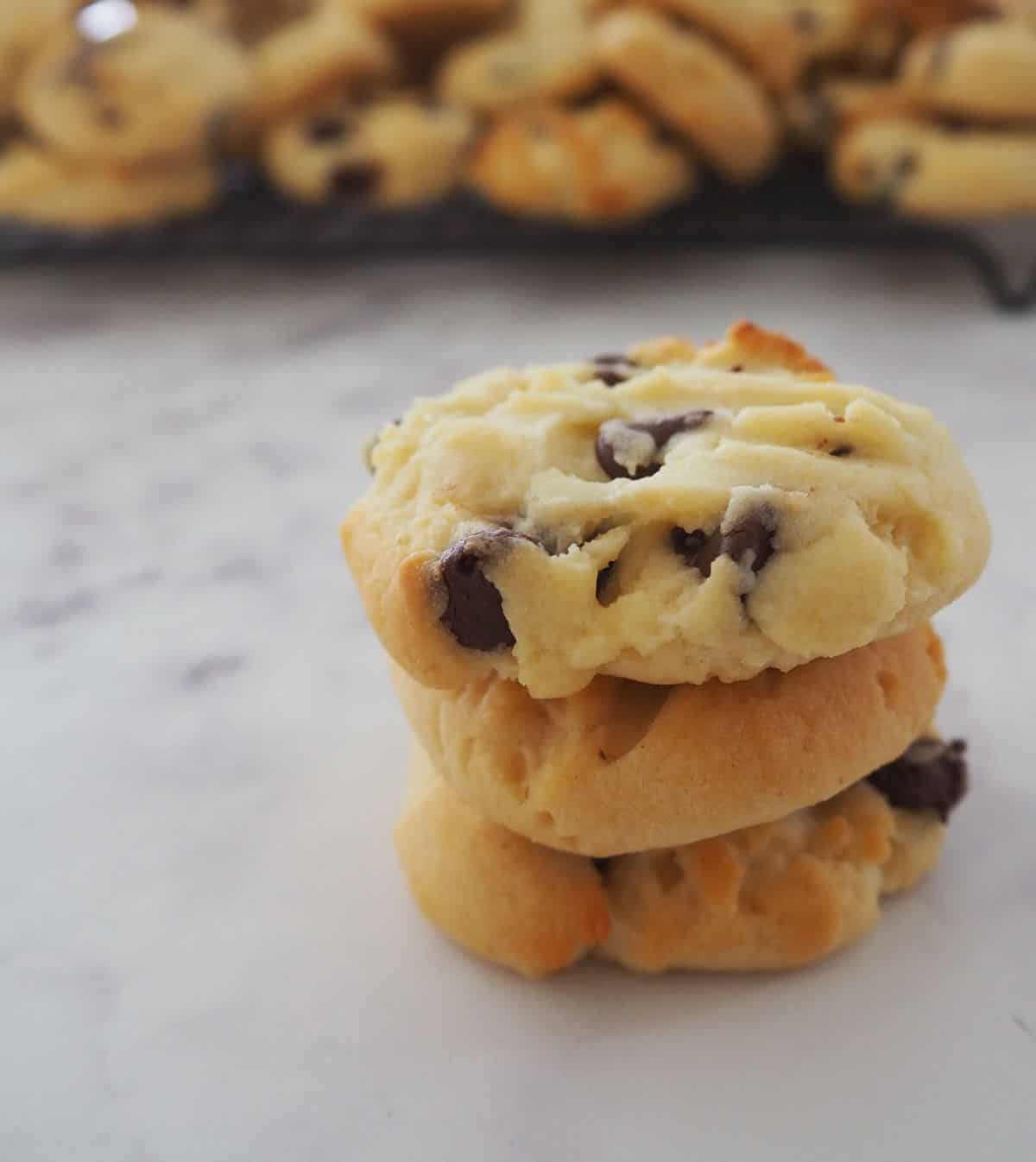 Three chocolate chip Condensed Milk Biscuits sitting on top of each other on a stone look bench top. There is a wire rack with more biscuits sitting on them behind.