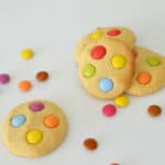 four smartie cookies on a white background with smarties sprinkled around them.