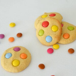 four smartie cookies on a white background with smarties sprinkled around them.