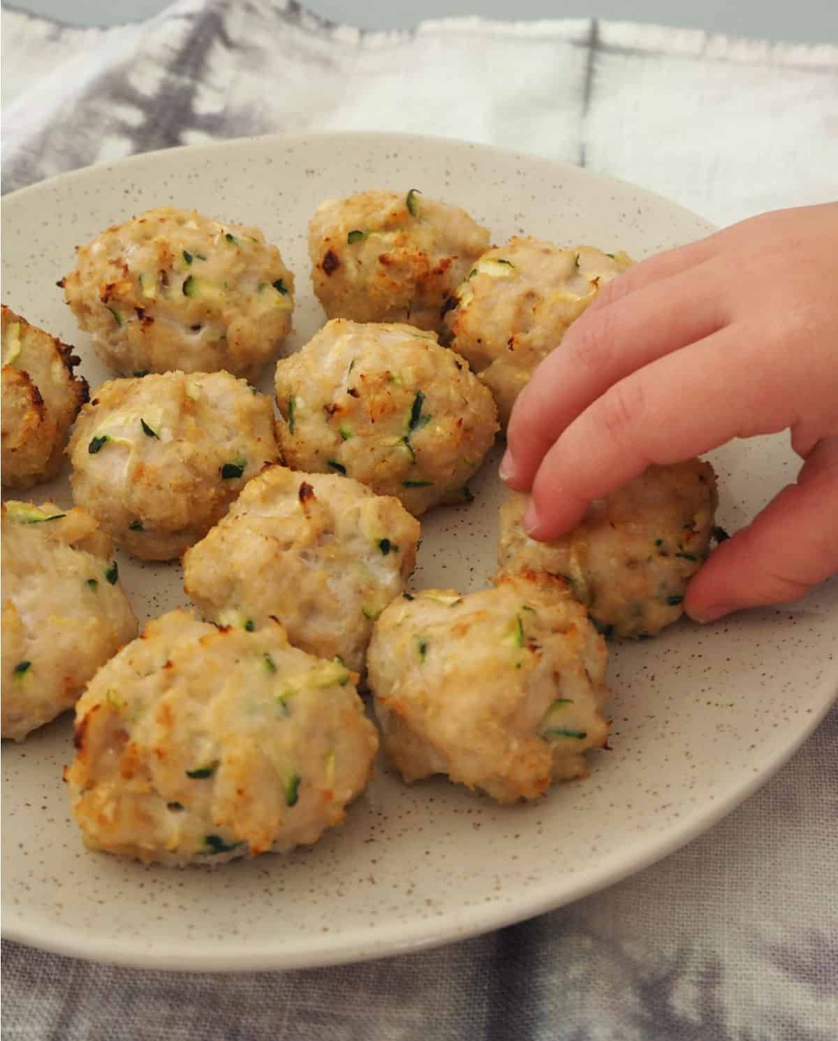 child's hand reaching for chicken zucchini and cheese ball on a white plate.