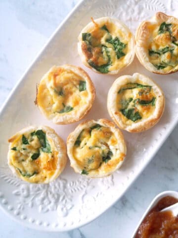 Overhead view of spinach and feta mini quiche on a white platter with a container of sauce in bottom right corner.