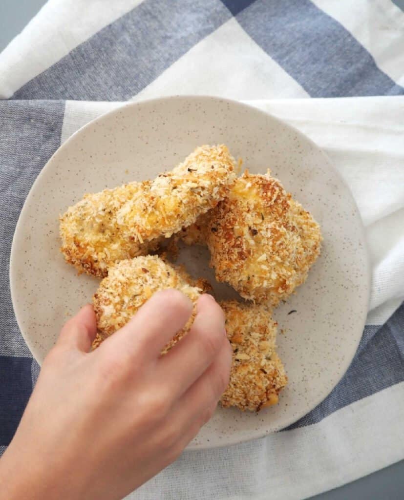 Child's hand reaching for a homemade chicken nugget on a speckled plate.