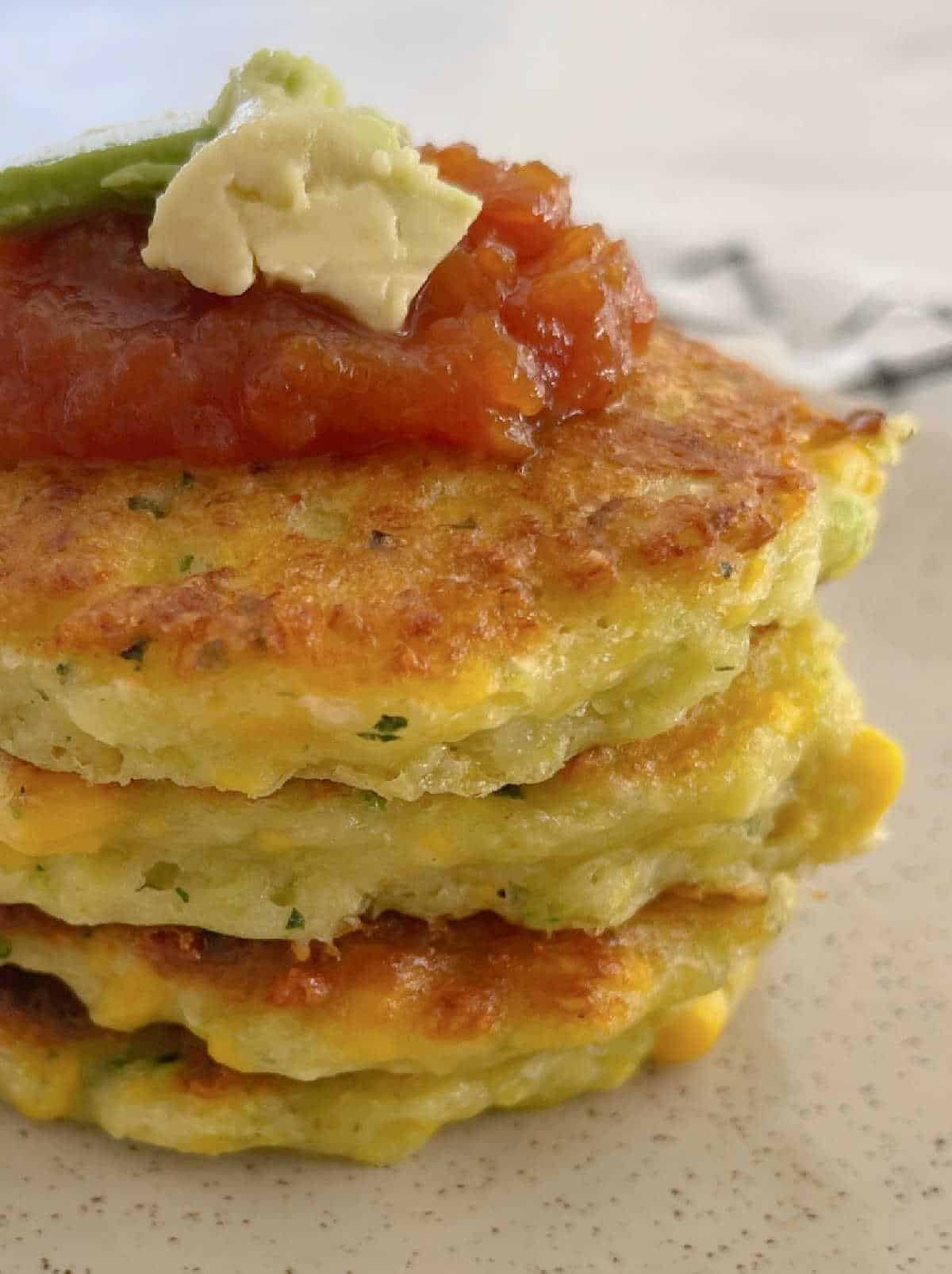 stack of corn and zucchini fritters on a speckled plate topped with tomato relish and avocado.