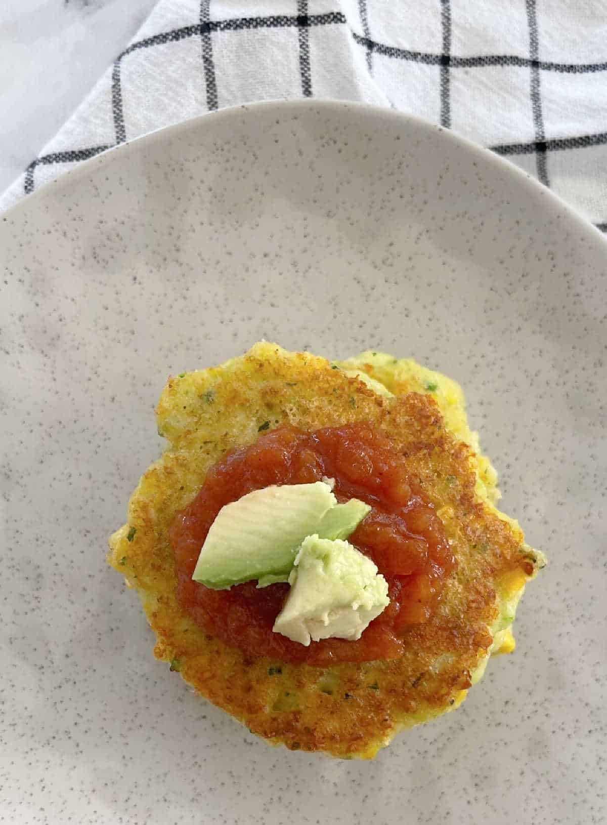 overhead of stack of corn and zucchini fritters on a speckled plate topped with tomato relish and avocado.