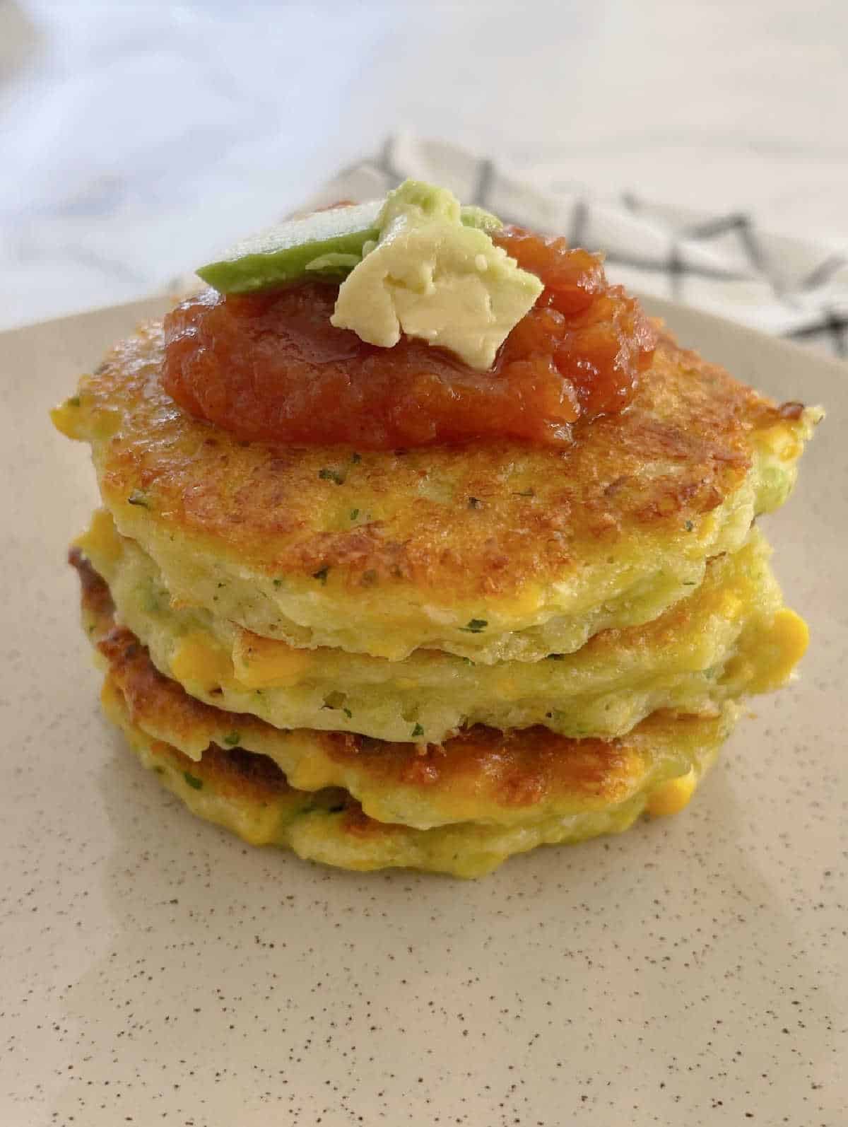 stack of corn and zucchini fritters on a speckled plate topped with tomato relish and avocado.