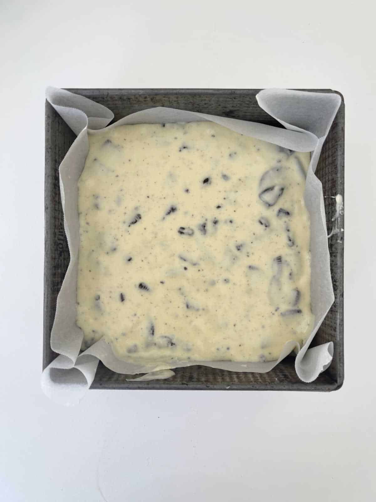 A square cake tin lined with baking paper that has white chocolate Oreo fudge mixture in it.