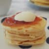 A stack of four pikelets sitting on a blue and white dotted plate topped with jam and cream.
