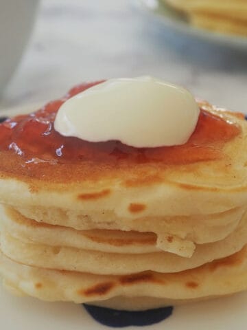 A stack of four pikelets sitting on a blue and white dotted plate topped with jam and cream.