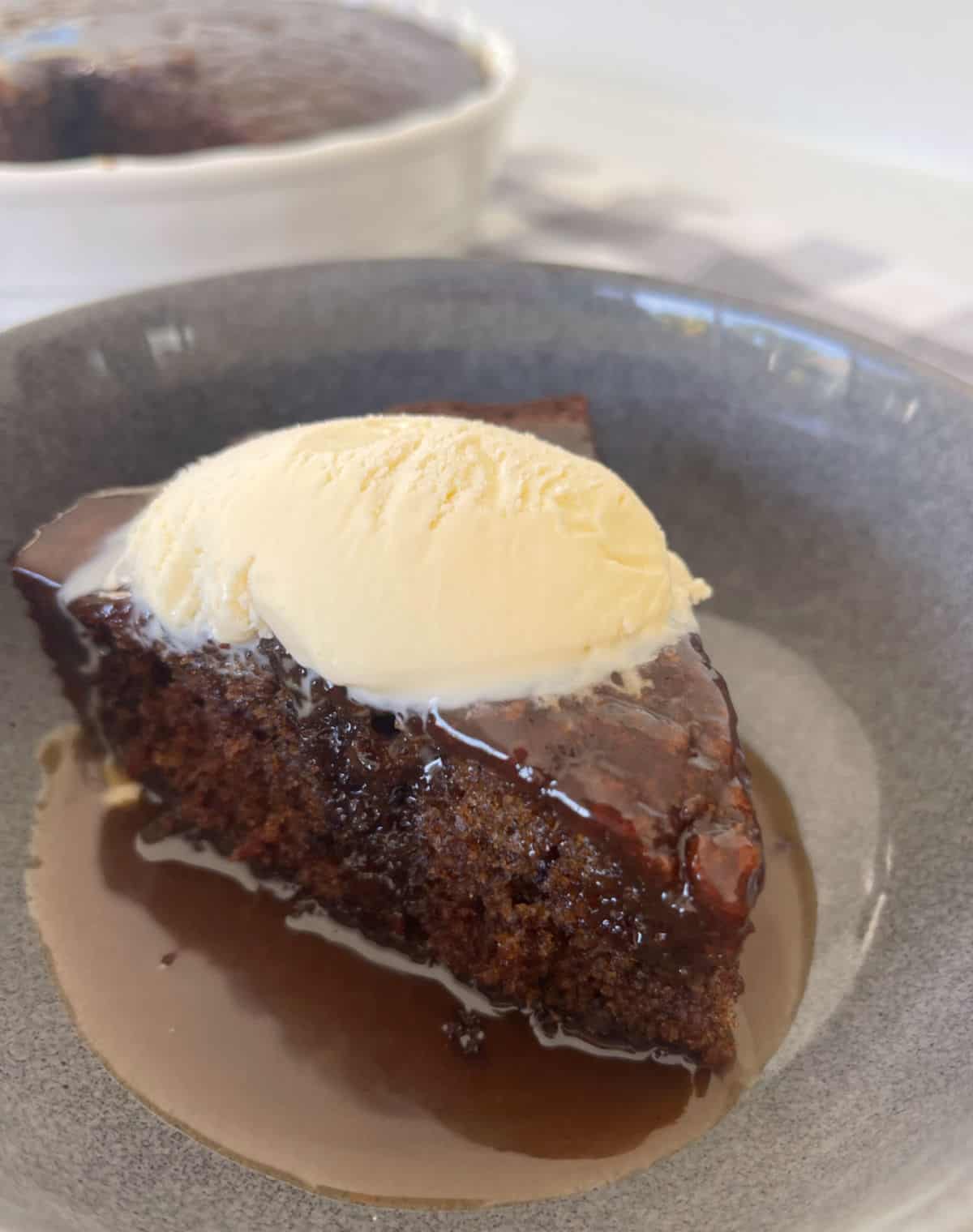 Piece of Sticky Date Pudding in a grey speckled bowl with caramel sauce and topped with ice cream.