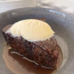 Piece of Sticky Date Pudding in a bowl with caramel sauce and topped with ice cream.