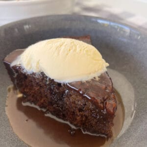 Piece of Sticky Date Pudding in a bowl with caramel sauce and topped with ice cream.
