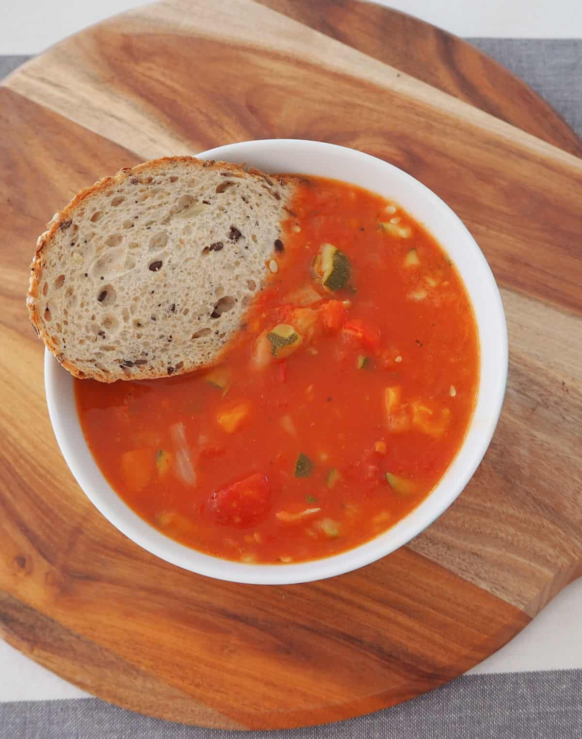 Bowl of vegetable soup with a piece of multigrain bread in it. Bowl is sitting on a wooden board.