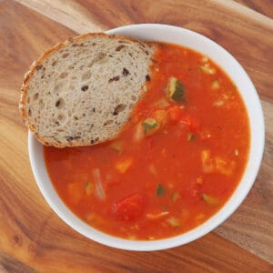 Bowl of vegetable soup with a piece of multigrain bread in it. Bowl is sitting on a wooden board.