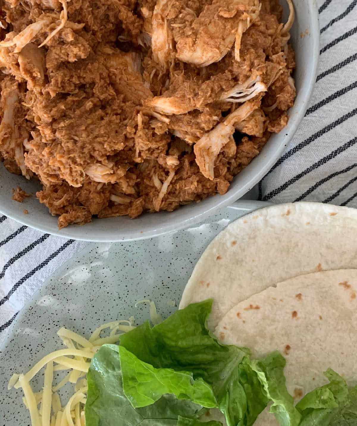 Shredded mexican chicken in a green speckled bowl next to a plate with tortillas, lettuce and cheese.