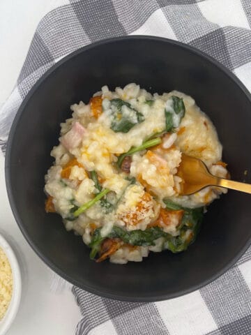 Bowl of Pumpkin Risotto sitting on a black and white check tea towel. A gold fork is sitting in the bowl with a container of parmesan cheese to the side.