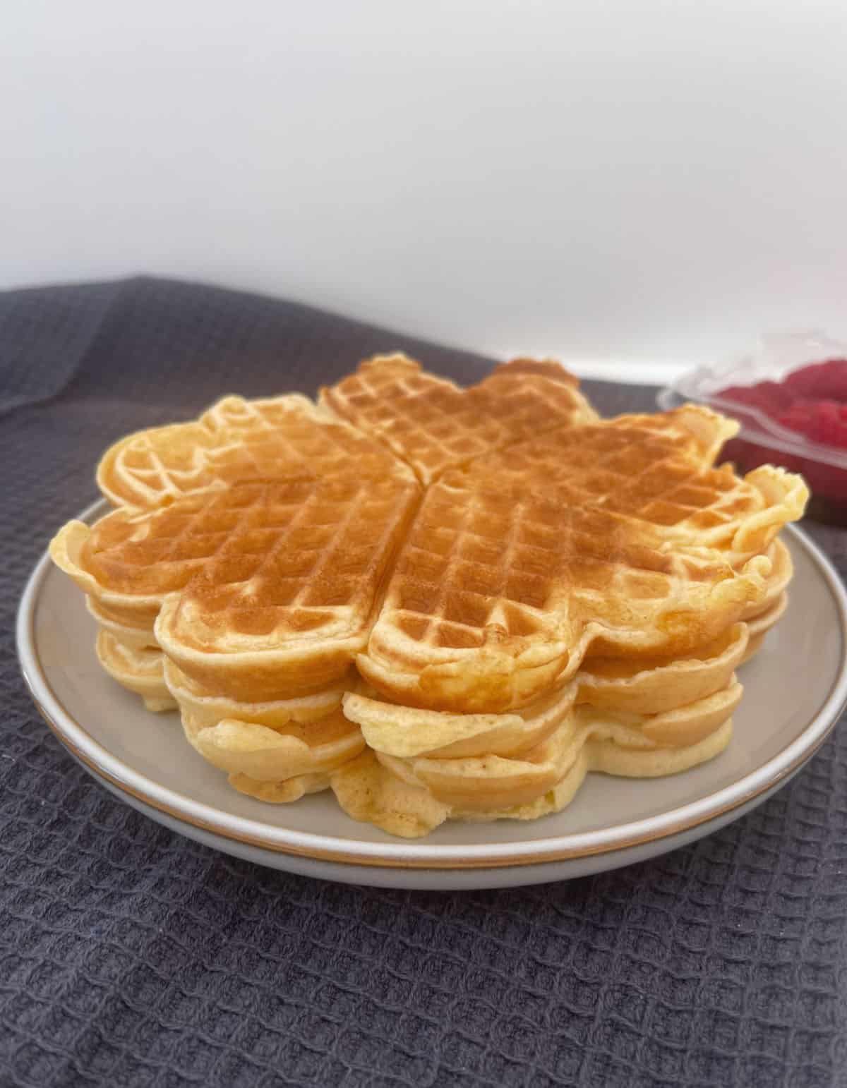 waffles stacked on top of each other on a grey plate with raspberries in the background.
