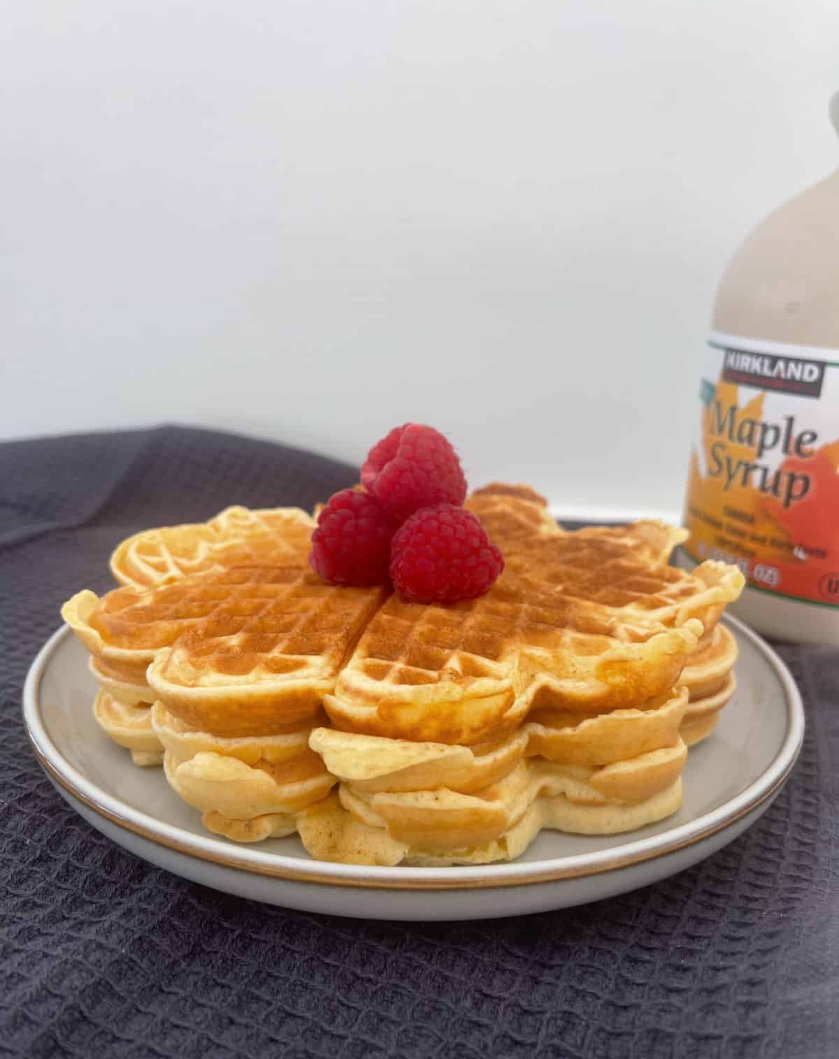A stack of waffles topped with raspberries and maple syrup in the background on a grey plate.