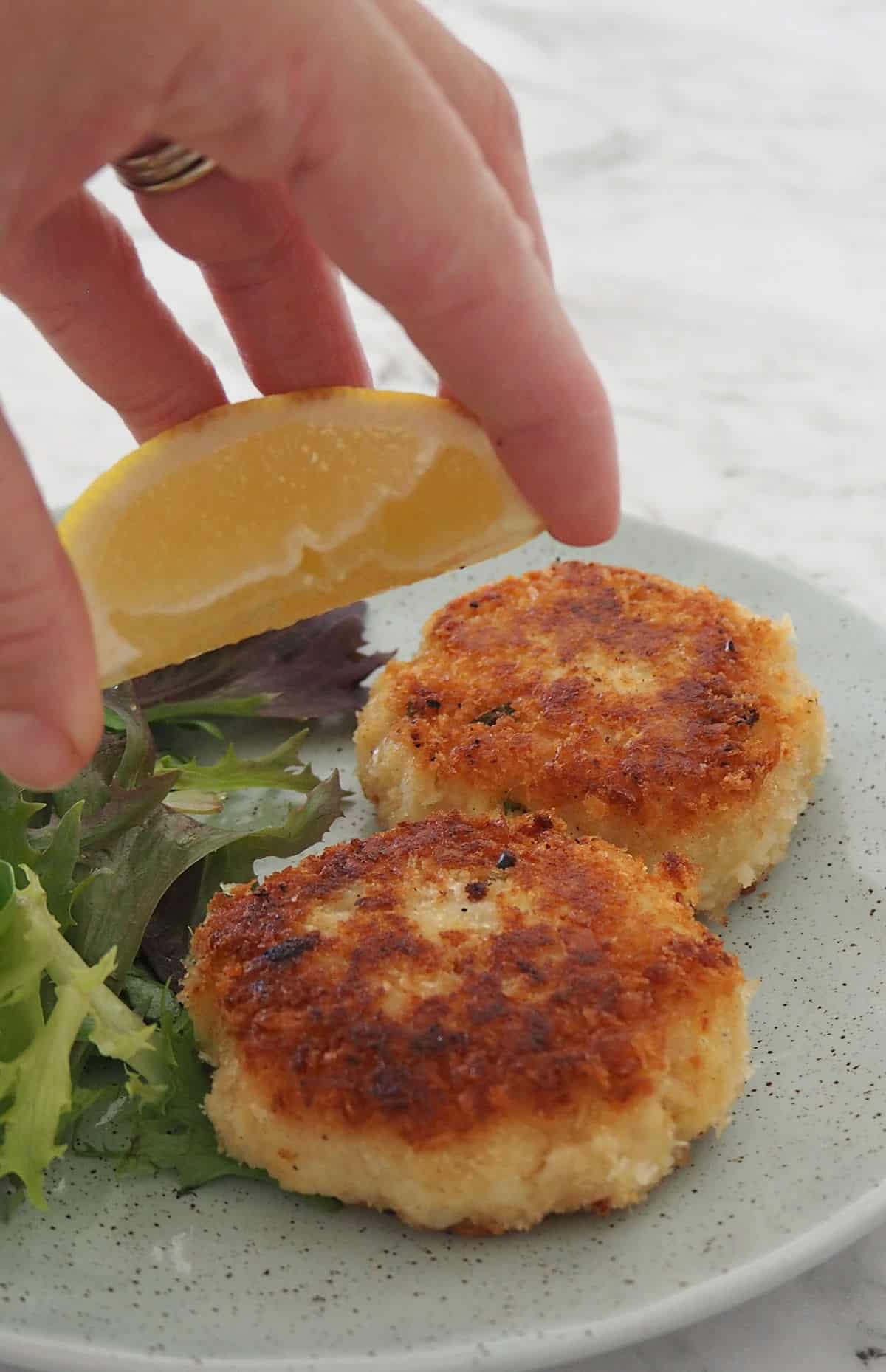 Adult squeezing lemon over two fish cakes on a green speckled plate.
