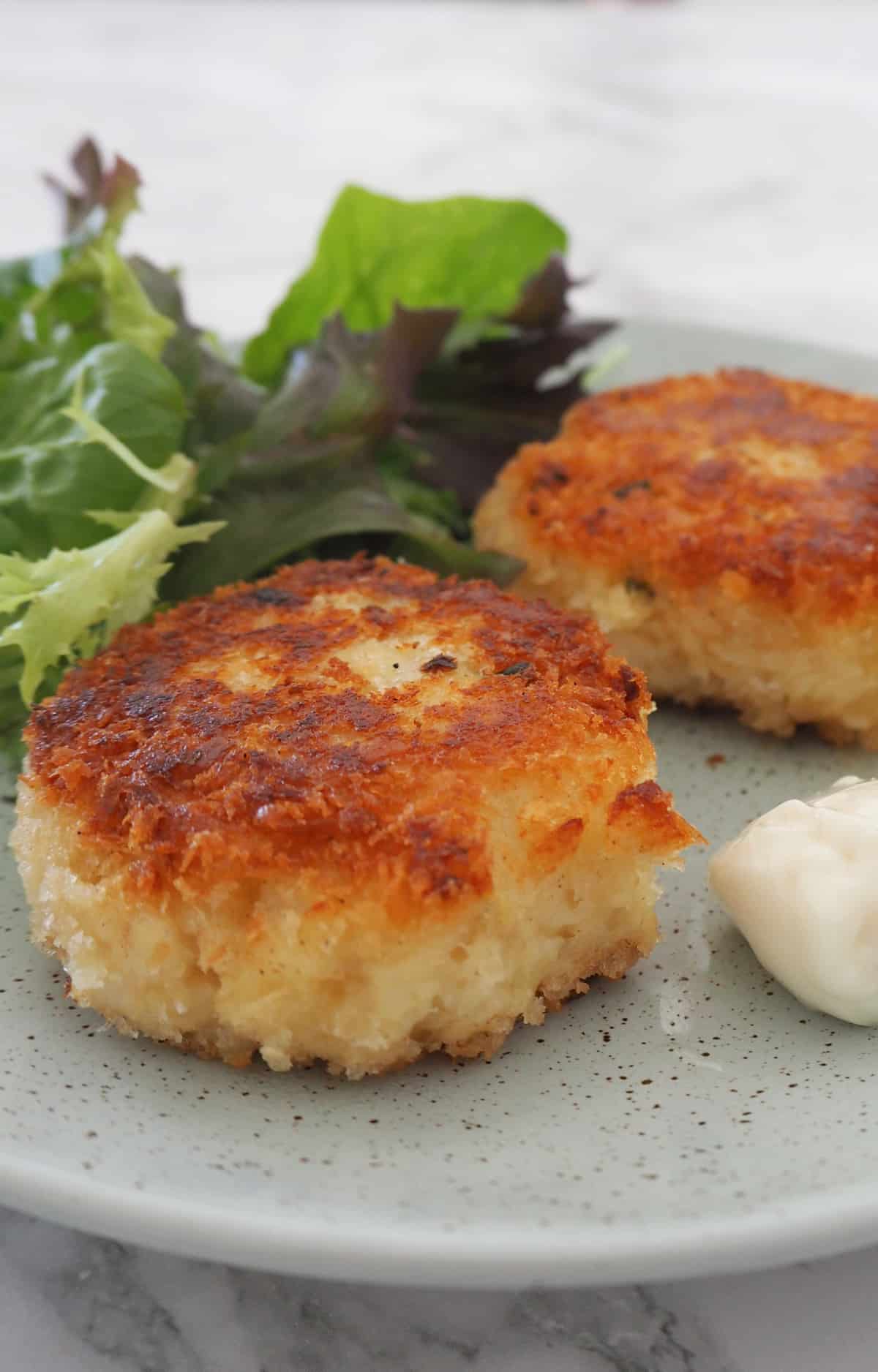 Two fish cakes on a green speckled plate. They are served with a green side salad and tartare sauce.