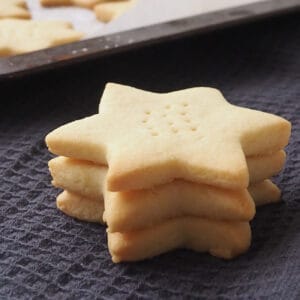 Three Shortbread Stars sitting on black towel with a tray of 3 ingredient shortbread behind them.