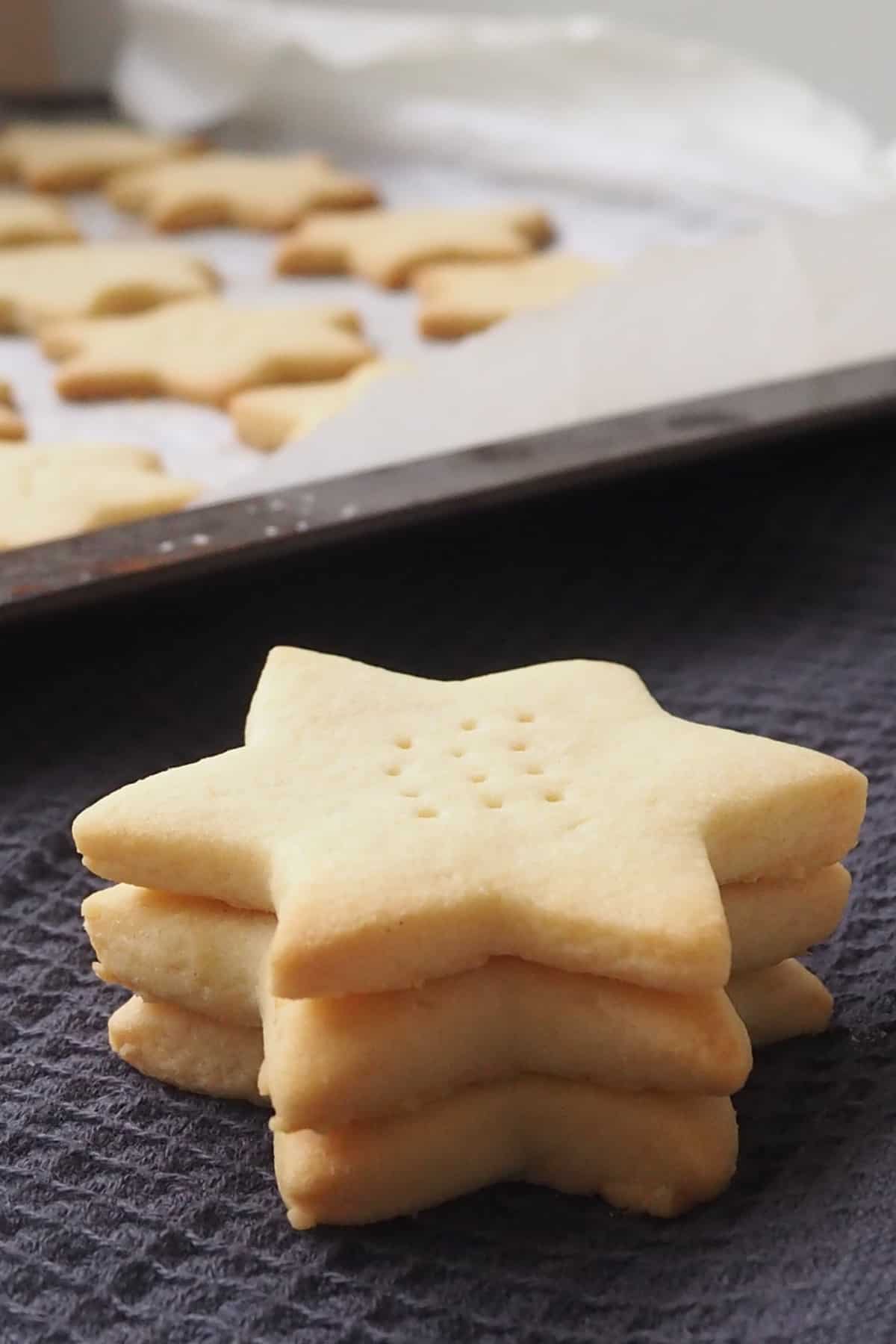 Three Shortbread Stars sitting on black towel with a tray of 3 ingredient shortbread behind them.