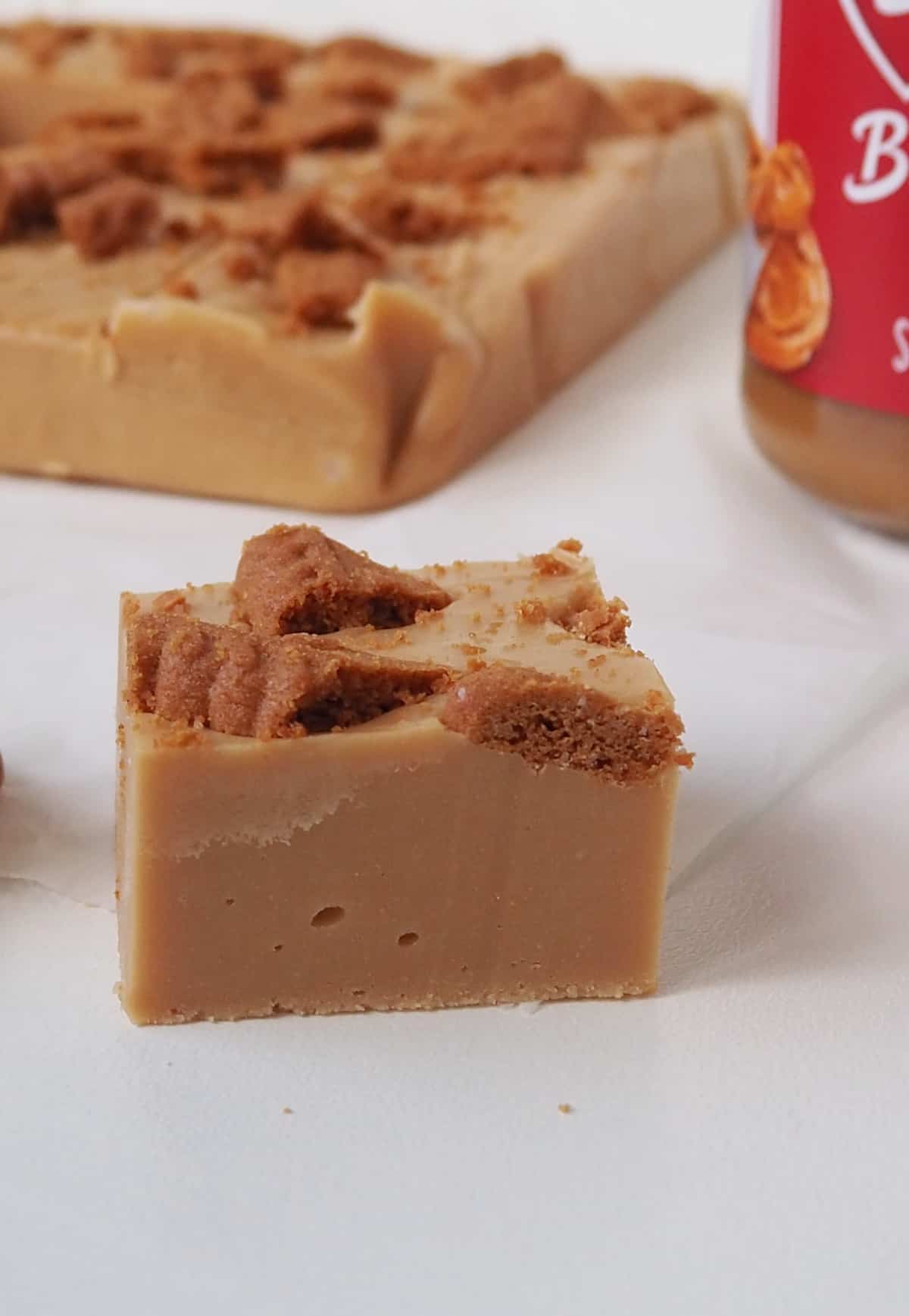 Close up of a piece of Biscoff Fudge on a sheet of baking paper. In the background is more fudge and jar of Biscoff spread.