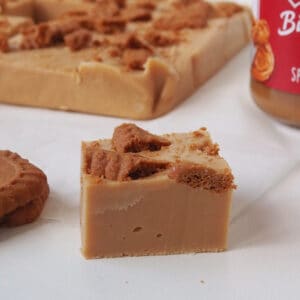 Close up of a piece of Biscoff Fudge on a sheet of baking paper. In the background is more fudge and jar of Biscoff spread.