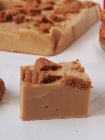 Close up of a piece of Biscoff Fudge on a sheet of baking paper. In the background is more fudge and jar of Biscoff spread.
