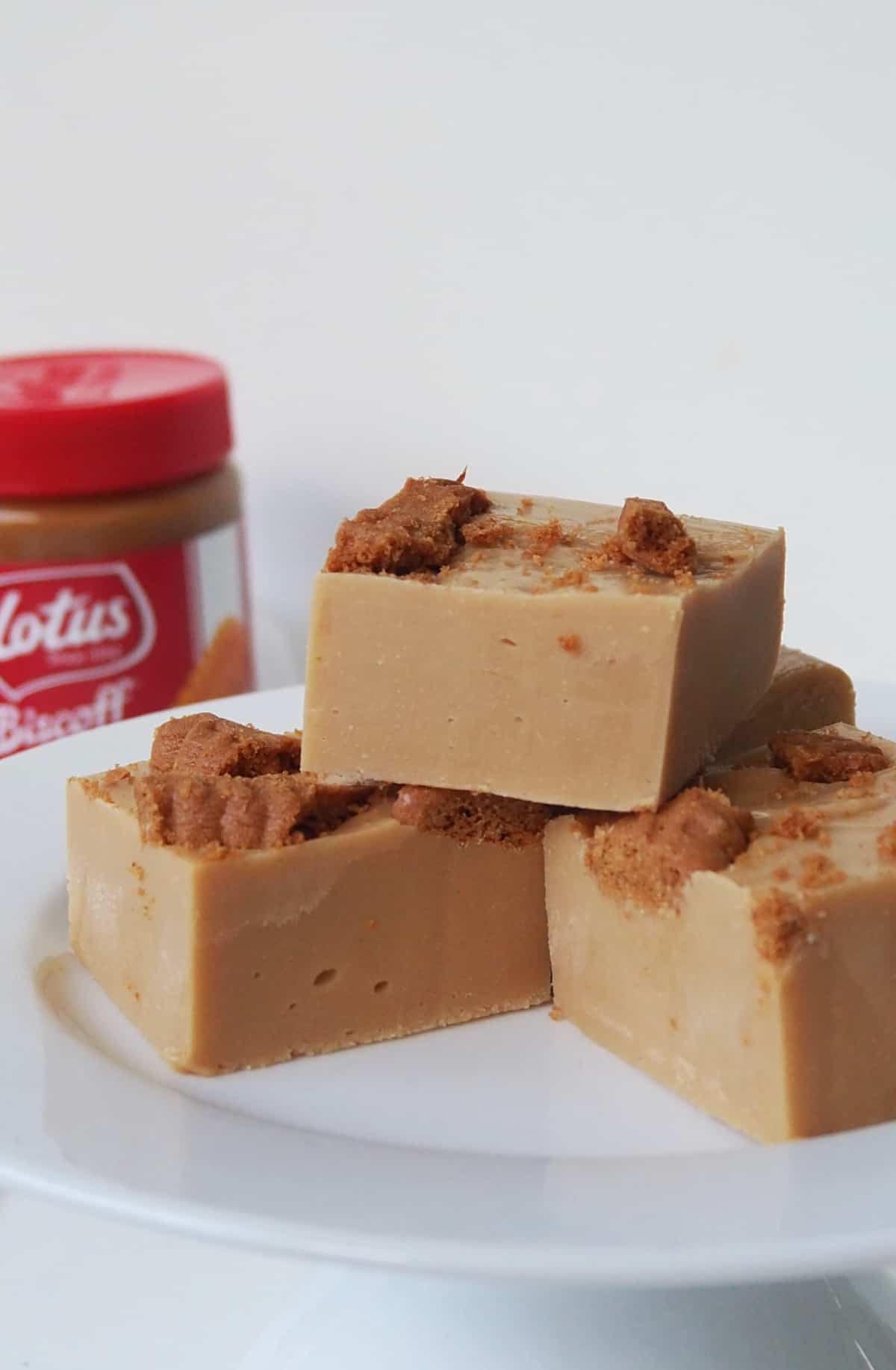 Three pieces of Biscoff Fudge sitting on a white plate. In the background is a jar of Biscoff spread.