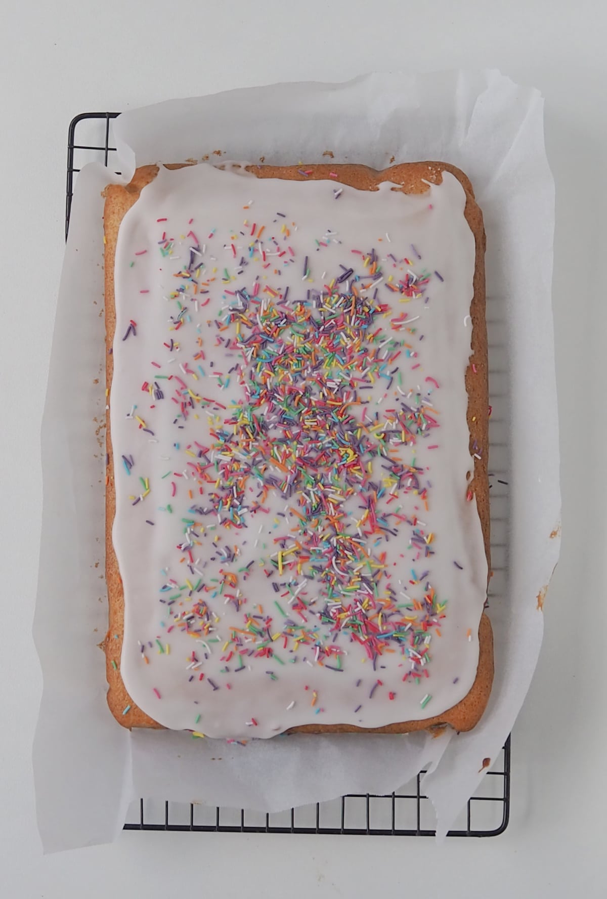 Overhead view of Vanilla Sheet Cake iced with a white frosting and decorated with sprinkles sitting on a sheet of baking paper on a wire rack.