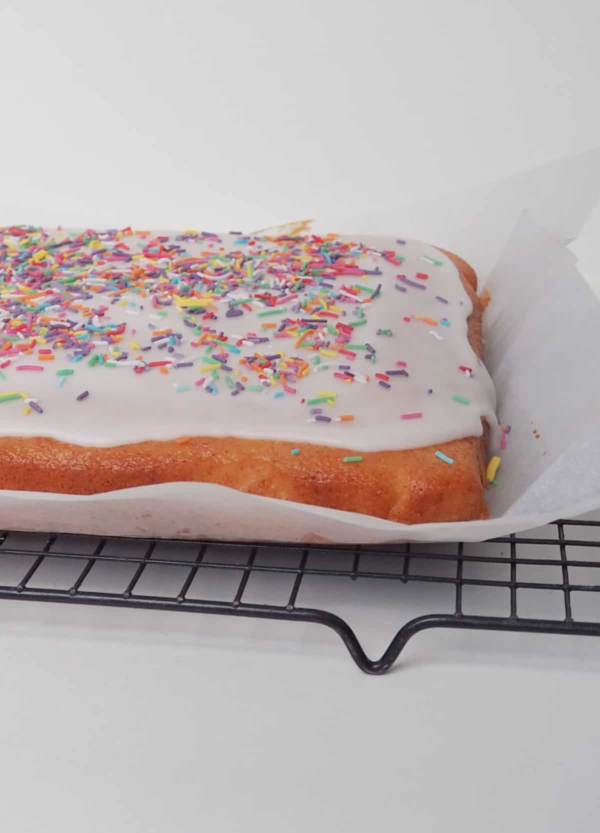 Vanilla Sheet Cake iced with a white frosting and decorated with sprinkles sitting on a sheet of baking paper on a wire rack.
