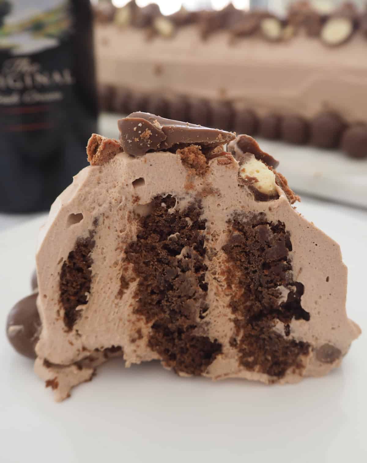 Side view of a piece of baileys chocolate ripple cake on a white plate. in the background is a bottle of Baileys Irish cream.