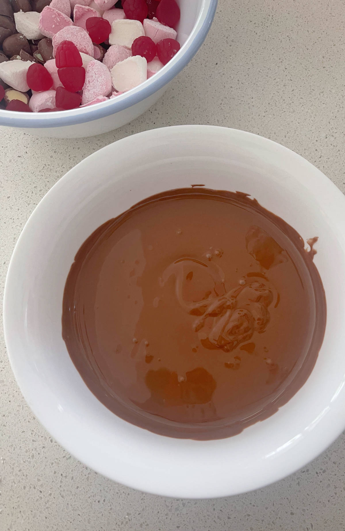 Melted milk chocolate in a white bowl. In the background is a bowl filled with ingredients to make clinkers rocky road.