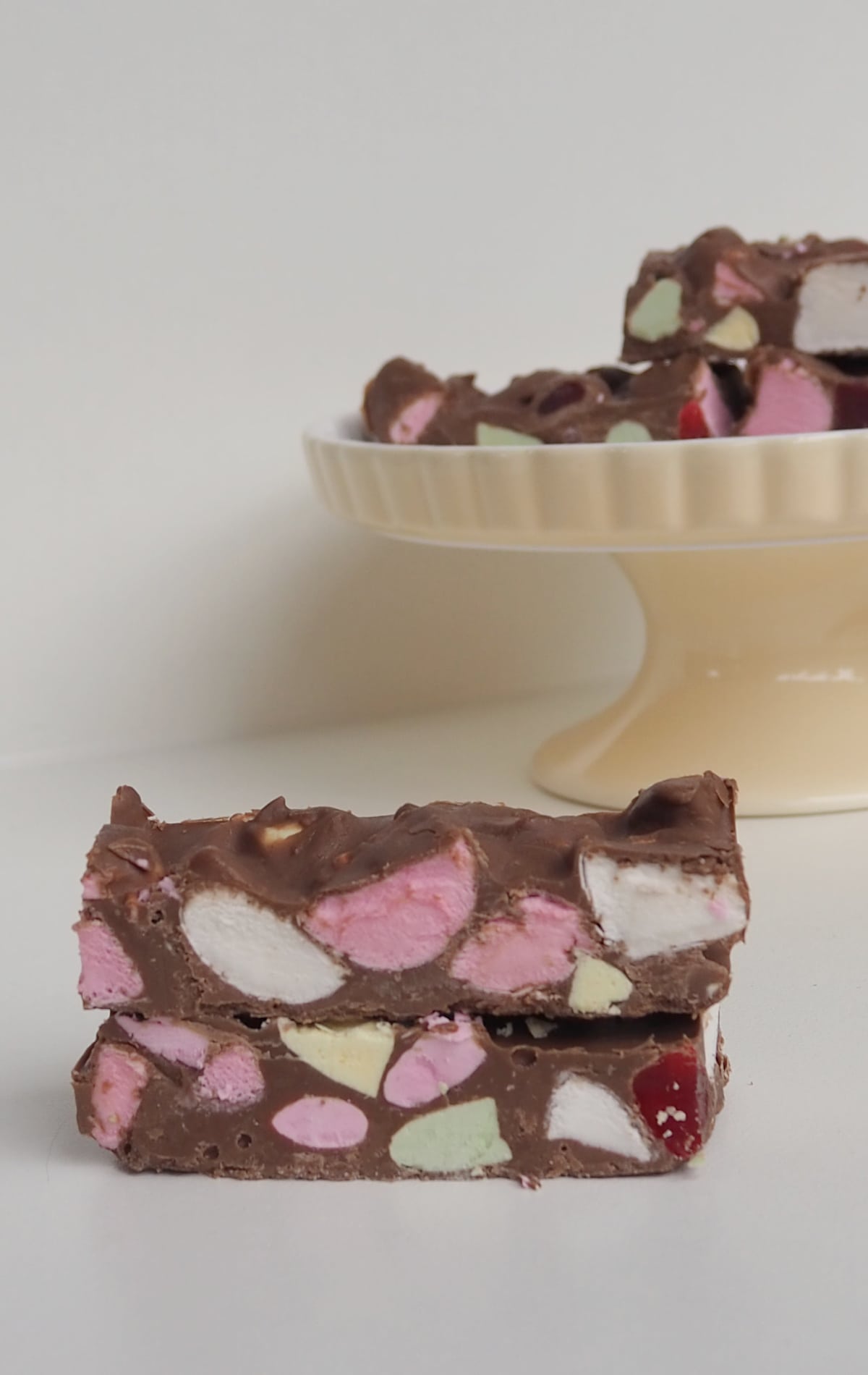 Two pieces of Clinkers Rocky Road stacked on top of each other. In the background is a cream cake stand with more pieces of rocky road on it.