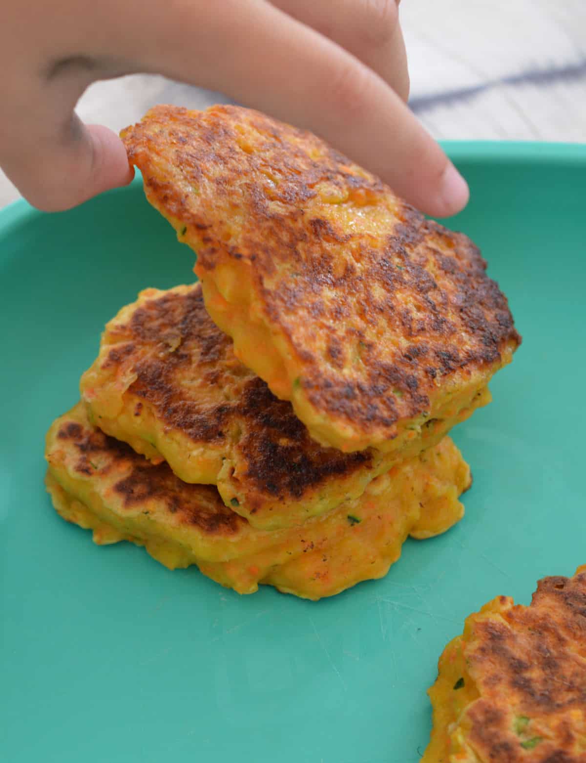 Child's hand reaching for a sweet potato fritter sitting on an aqua plate.