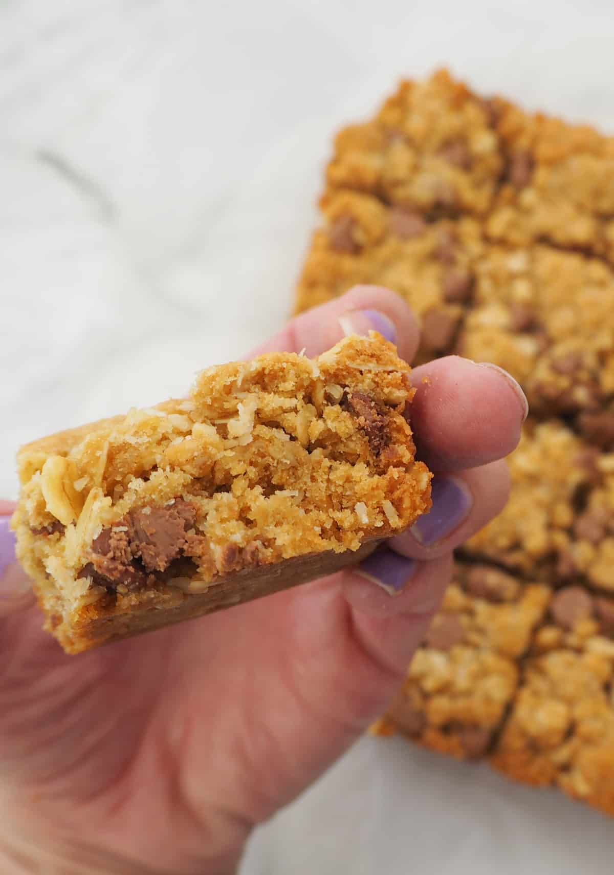 adult holding a piece of chocolate chip Anzac slice.