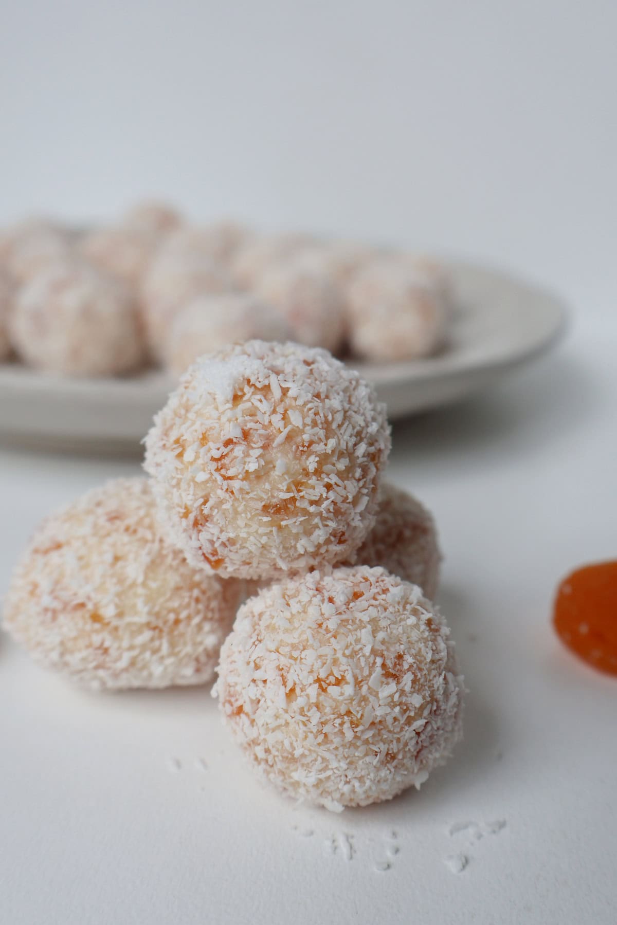 Four apricot and coconut stacked. Behind them is a dried apricot and plate with the rest of the balls. 