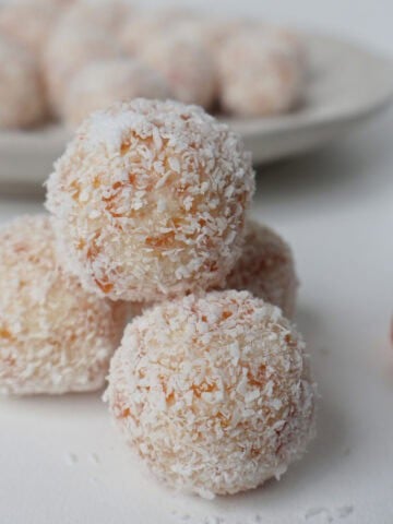 Four apricot and coconut stacked. Behind them is a dried apricot and plate with the rest of the balls.