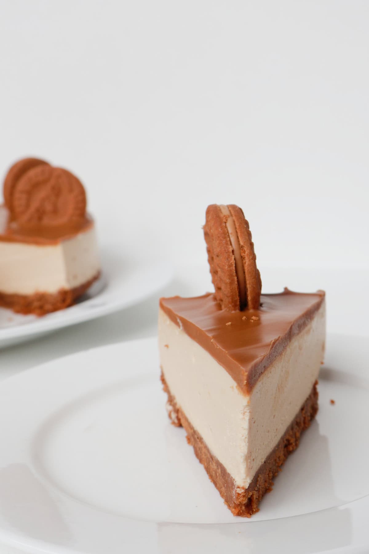 Front view of Slice of Biscoff Cheesecake on a white plate. In the background is the cheesecake on a white platter.