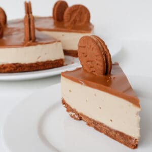 Slice of Biscoff Cheesecake on a white plate. In the background is the cheesecake on a white platter.